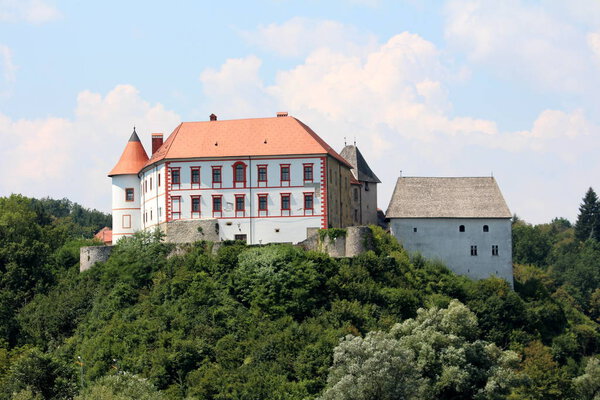 Medieval castle with completely renovated front walls, guard tower, roof tiles and surrounding stone walls located on top of the hill surrounded with dense trees and forest vegetation with cloudy blue sky in background