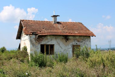 Küçük terkedilmiş kırmızı tuğla ev ile harap beyaz cephe, yok çatı karoları, kapılar ve pencereleri tamamen yüksek çim ve arka planda bulutlu mavi gökyüzü ile diğer bitki örtüsü ile çevrili