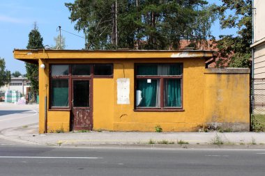 Arka planda büyük ağaçlar ve inşaat malzemesi ile asfalt yolun önünde kırık ahşap kapılar ve cephe ile İşyeri binaya terkedilmiş eski harap sarı güvenlik görevlisi girişi