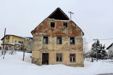 Kırık pencereleri ile terkedilmiş eski harap ev, duvarlar gelen cephe ve tuğla düşmüş, derin kar döşeli yolun yanında ve soğuk kış gününde arka planda modern banliyö evi ile kaplı