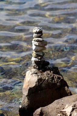 Beach stones stacked on one straight pile on top of large rock used for meditation or as artistic installation with clear sea in background on warm sunny day