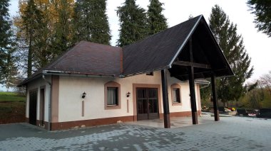 Mezarlık ve yüksek ağaçların yanında büyük kapılar, iki pencere, ışıklar ve beton fayans bahçe ile mezarlık morga ön girişi