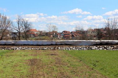 Uzun sel koruma duvarı, arka planda bulutlu mavi gökyüzü ile uzun ağaçlar ile çevrili banliyö aile evleri ve küçük binalar koruyan beyaz jeotekstil kumaş ile kaplı kutu bariyerleri yapılmış