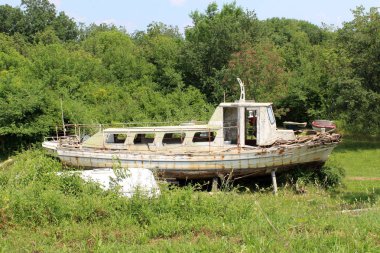 Kırık pencereler, kapılar, ahşap tahtalar ve paslanmış metal parçalar ile terkedilmiş eski harap ahşap tekne, sıcak güneşli günde kesilmemiş çim ve orman bitki örtüsü arasında arka bahçede sol