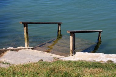 Çim ve beton nehir bankasında korkuluklar içeren, sıcak güneşli bahar gününde doğrudan suya giden kısmen batık ahşap adımlar