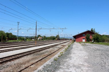 Büyük kısmen paslanmış elektrik direkleri ve arka planda eski ve yeni tren kompozisyonlar altında birden fazla demiryolu parçaları ile demiryolu istasyonu girişi sıcak yaz aylarında ağaçlar ve diğer bitki örtüsü ile kaplı küçük binanın yanında kalkış bekliyor