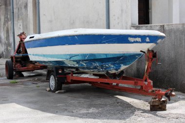 Taş duvarın önüne park edilen kırmızı metal römork üzerinde onarım ihtiyacı olan küçük beyaz ve mavi tekne