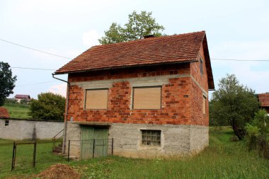 Kapalı pencere Jaluzi ve günbatımında arka planda kesilmemiş çim ve açık mavi gökyüzü ile çevrili harap ahşap garaj kapıları ile tamamlanmamış terk kırmızı tuğla aile evi