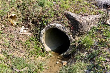 Akan su ile hasarlı beton Drenaj Boru çıkışı ve kir ile çevrili, taş, kesilmemiş çim, kurutulmuş yaprakları ve küçük hasarlı plastik boru