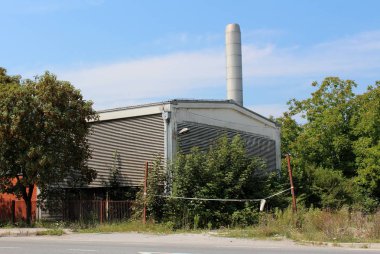Old abandoned factory with red brick wall and metal insulation, rusted fence without doors and asphalt road in front surrounded with large trees, overgrown grass and other vegetation with tall metal chimney and cloudy blue sky in background