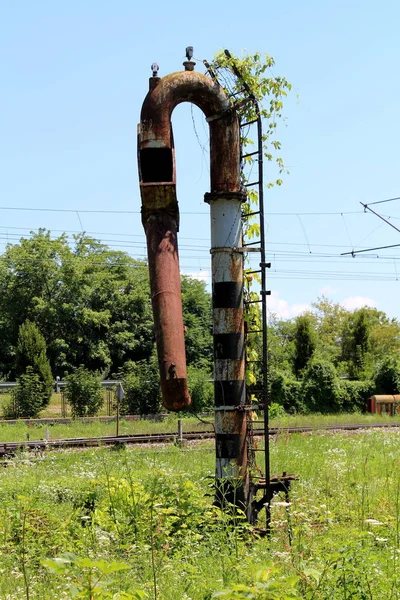 Rusted metal railway white and black water pump with rusted and overgrown metal ladder mounted on side surrounded with high uncut grass with trees and clear blue sky in background
