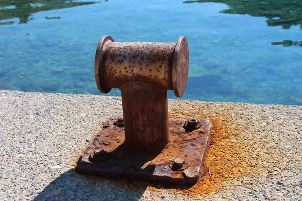 Rusted strong old iron mooring bollards screwed on local concrete and stone pier with four rusted metal bolts and with calm blue sea in background