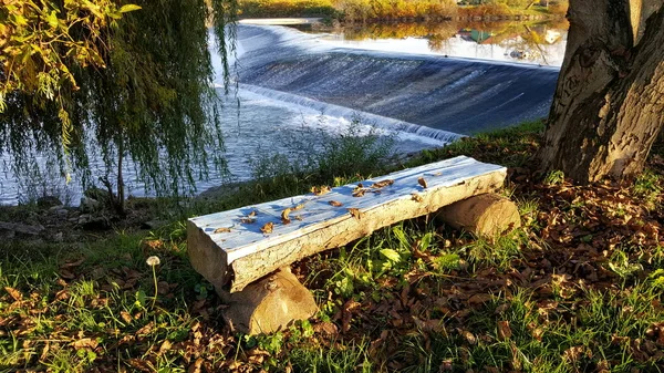Wooden bench made from small pieces of cut trees in front of river waterfall and surrounded with trees, leaves, uncut grass and single flower