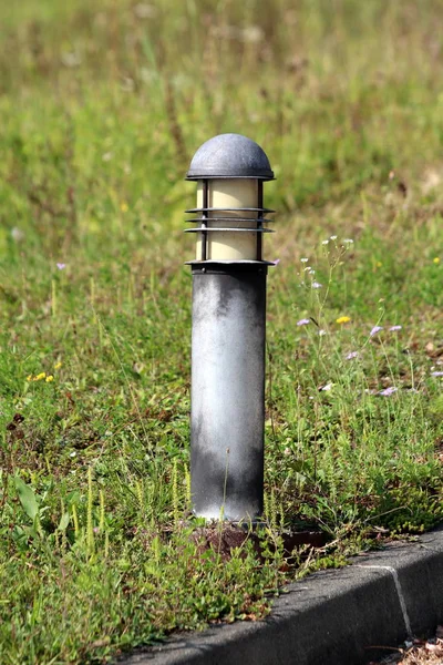 Modern style outdoor lamp with faded black color next to paved driveway surrounded with high uncut grass and small flowers on warm summer day