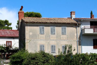 Kapalı harap ahşap pencere Jaluzi ve sıcak güneşli gün arka planda açık mavi gökyüzü ile diğer evler ve yoğun ağaçlar ile çevrili paslı baca ile büyük eski taş Akdeniz evi