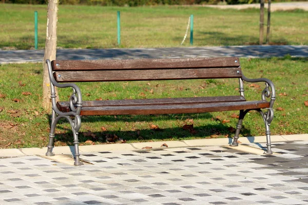 木の板とバロック様式を使用している公共の公園のベンチは 背景に草や落ち葉と黒と灰色の石のタイルに取り付けられた鉄のサポート — ストック写真