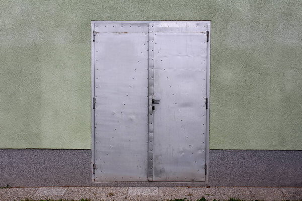 Grey sheet metal doors with small door handle mounted on house wall with stone tiles in front on warm sunny day