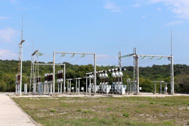 Açık mavi gökyüzü ile çakıl ve orman ile çevrili elektrik telleri ile bağlı koyu kahverengi seramik ve cam izolatörleri ile çeşitli cihazlardan yapılmış güçlü metal desteği üzerinde yerel güç santrali yoğun destek elektrik donanımları, arka
