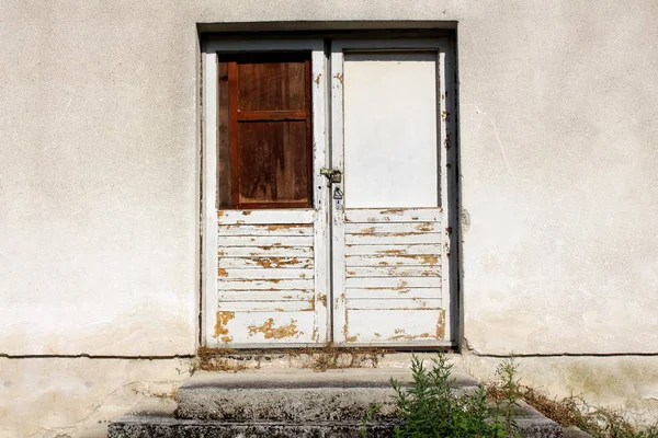 Locked white wooden doors with cracked dilapidated paint, broken windows replaced with boards and rusted padlock on old grey wall with concrete steps in front on warm sunny day