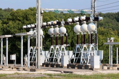 Sıcak güneşli bir gün arka planda çakıl ve orman ile çevrili elektrik teller ile bağlı koyu kahverengi seramik ve cam izolatörler için güçlü metal desteği ile yerel güç santralinde elektrik donanımları yoğun dizi