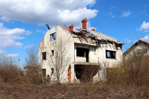 Damaged family house during war with missing windows and completely destroyed roof with only chimneys left untouched surrounded with overgrown underbrush and cloudy blue sky background