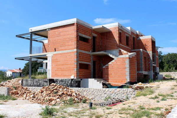 Sıcak güneşli bir gün açık mavi gökyüzü arka planda taşlar ve yapı malzemesi ile çevrili inşaat altında modern büyük kırmızı tuğla aile evi