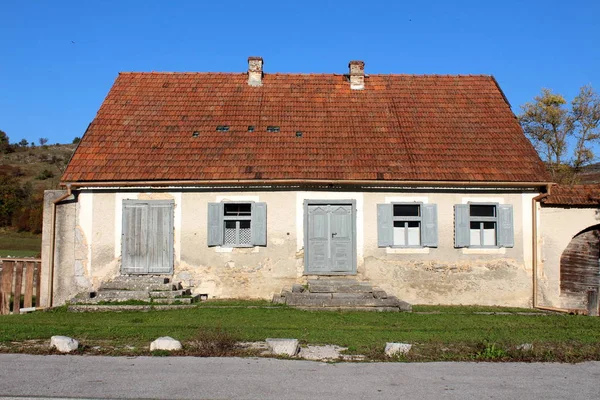 Old abandoned traditional family house with dilapidated facade and wooden window blinds surrounded with grass and clear blue sky in background on warm sunny day