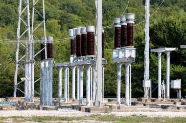 Uzun boylu koyu kahverengi seramik izolatörler güçlü metal borular üzerine monte ve sıcak güneşli gün arka planda çakıl ve yoğun orman ile çevrili yerel santralin içinde elektrik telleri ile bağlı