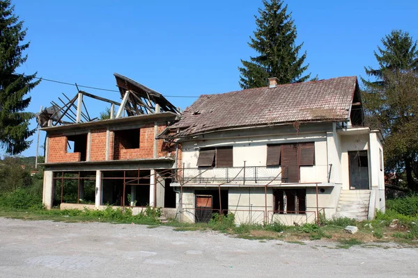 Attached family houses destroyed in war abandoned by owners with missing windows and broken roof surrounded with uncut grass and gravel in front and tall trees with clear blue sky in background on warm sunny day