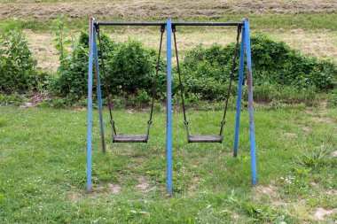 Outdoor public playground equipment metal swing with partially rusted blue frame and dilapidated wooden seats surrounded with uncut grass and other small forest vegetation in background