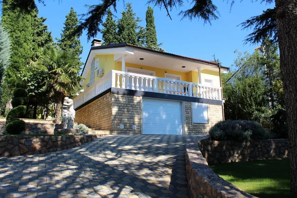 Modern Mediterranean stone and concrete house with stone driveway, large trees, small plants and sculptures in its surroundings