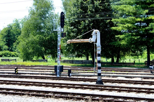 Old metal railway white and black water pump next to railway tracks and traffic light mounted on strong metal pole and surrounded with gravel, tracks and signaling posts with trees and concrete fence in background