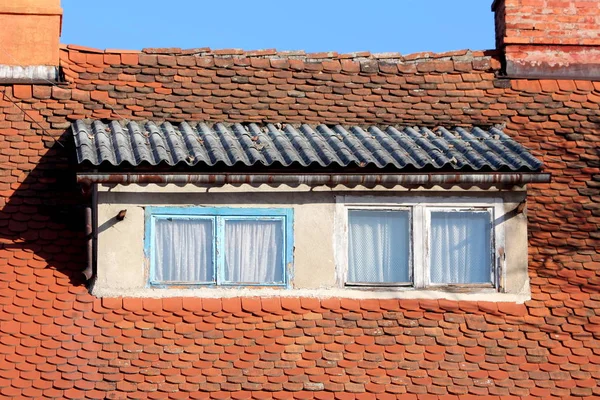 Roof windows with light blue and white dilapidated wooden frames surrounded with small roof tiles and rusted gutter next to two red brick chimneys on clear blue sky background