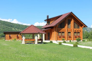 Taş çardak ve ahşap günlük aracı ile ahşap günlük kabin evi taş giriş, arka planda mavi gökyüzü ile küçük çiçekler ve orman dolu kesilmemiş çim çevrili döken