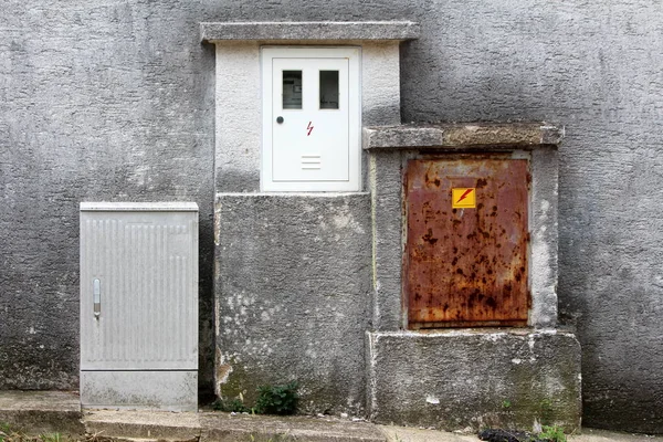 New electrical meter box with single meter mounted on dilapidated wall next to rusted old metal and new plastic grey electrical boxes next to concrete sidewalk on warm sunny day