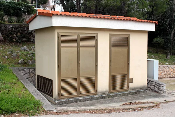 Small local electricity substation building with metal doors surrounded with traditional stone wall, grass, trees and other vegetation