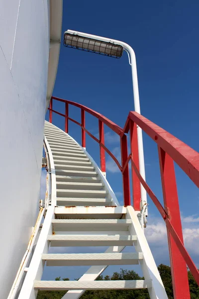 Spiral metal stairs mounted on side of large storage silo with red security fence and large light on clear blue sky background on warm sunny day