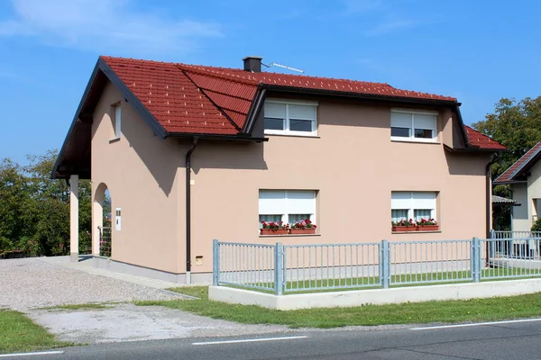 Yeni cephe ile tamamen yenilenmiş küçük banliyö aile evi, taze çiçekler ile beyaz pencereler, yeni kiremit ve evye, gri metal çit ile çevrili, kesilmemiş çim ve arka planda ağaçlar ve açık mavi gökyüzü ile çakıl Garaj girişi