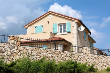 Yeni cephe ve arka planda yoğun hedge ve bulutlu mavi gökyüzü ile çevrili açık mavi ahşap pencere jaluzi ile modern Akdeniz Villa önünde üst Ferforje çit ile geleneksel taş duvar