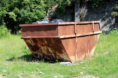 Tamamen paslanmış eski büyük açık çöp çöp kutusu sıcak güneşli gün arka planda kesilmemiş çim ve orman bitki örtüsü ile çevrili çöp dolu