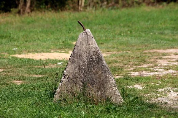 Heavy pyramid like concrete barrier with strong rusted metal hook on top surrounded with high uncut grass and trees in background