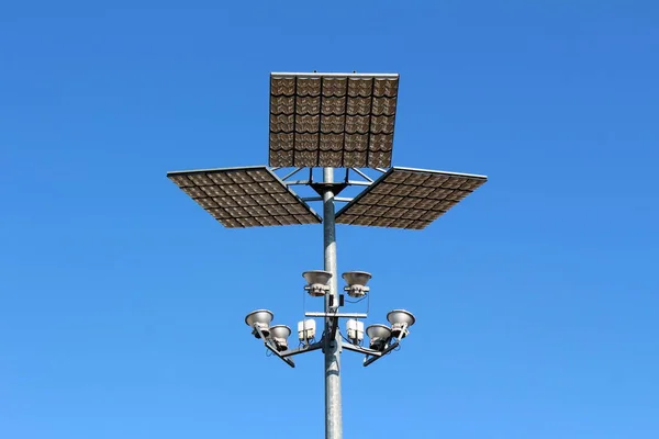Light grey strong metal pole holding modern LED street light reflectors pointed towards large reflective panels on clear blue sky background