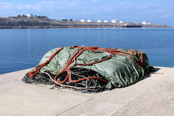 Pile of fishing nets covered with nylon protection during winter on concrete pier edge surrounded with calm sea and industrial complex with oil tanker in background