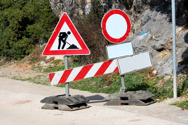 Road closed under construction metal road signs mounted on plastic holders on paved road with rocks and trees in background