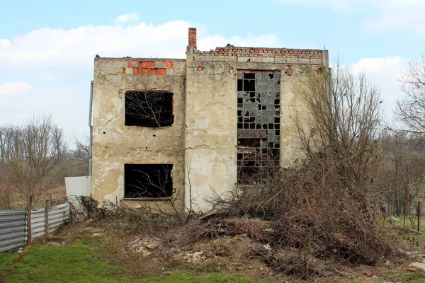 Ruined house with damaged facade and without roof surrounded with overgrown dried vegetation and garbage on cold winter day