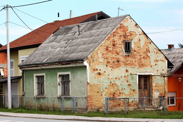 Shrapnel damaged small family house during war now abandoned by owners surrounded with uncut grass and broken partially rusted fence on warm sunny day