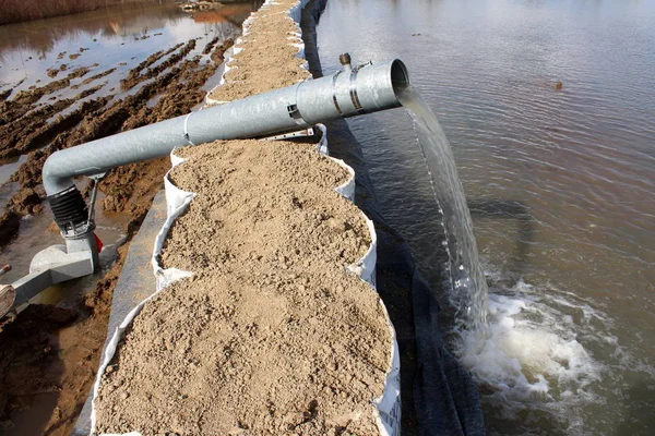 Water pump pumping flood water over tall sandbox barriers through large metal hose on sunny winter day