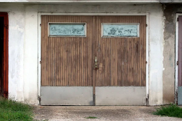Wooden garage doors made of dilapidated wooden boards with painted windows and metal plate rain protection mounted on cracked wall with old asphalt driveway surrounded with tall uncut grass