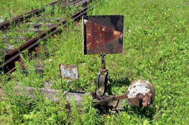 Sıcak güneşli bir gün küçük çiçekler ile büyümüş kesilmemiş yeşil çim ile çevrili demiryolu pistin yanında ahşap tahta monte metal işareti ile paslanmış Metal siyah ve beyaz demiryolu anahtarı mekanizması
