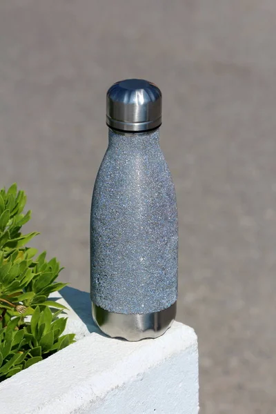 Shiny metal thermos drinking bottle with partially rough texture left on edge of white concrete flower pot on public sidewalk on warm sunny day